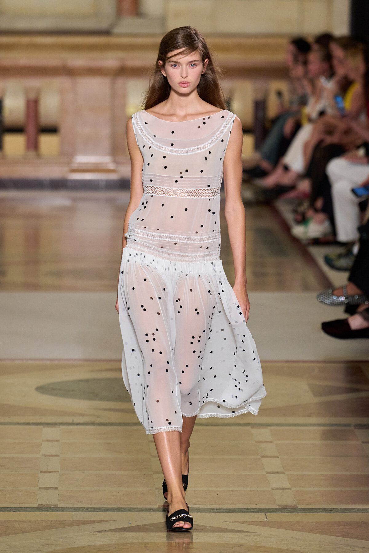 Model walks the runway in a sheer white polka-dot dress with lace details and a gathered hem, paired with black sandals.