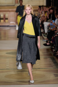 Model walking the runway in a yellow sleeveless top, black leather coat, and gray textured midi skirt with a belt; white flats and a silver handbag in hand.