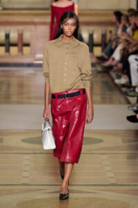 Model walking a runway in a tan button-down sweater and red leather culottes with a black belt, carrying a white handbag.