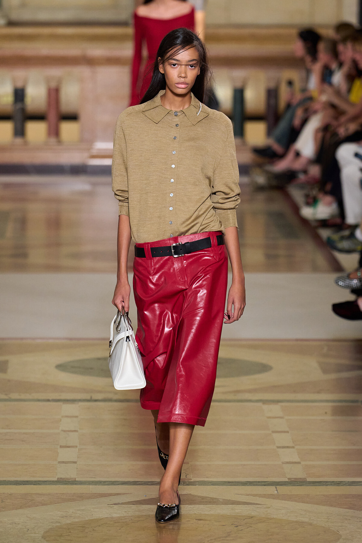 Model walking a runway in a tan button-down sweater and red leather culottes with a black belt, carrying a white handbag.