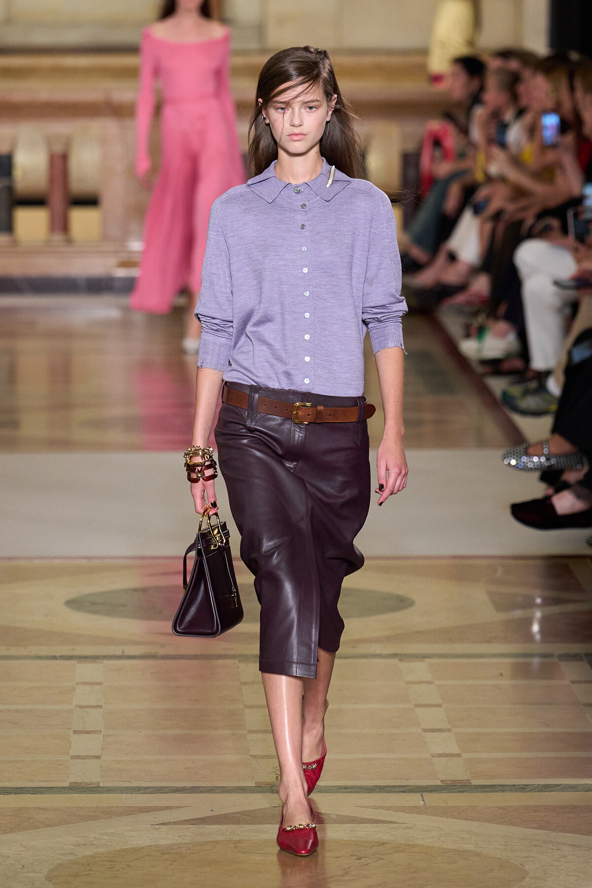 Model walking a fashion runway in a lavender button-down top, brown belt, burgundy leather midi skirt, red heels, and a small black handbag.