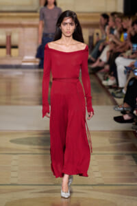 Model walking the runway in a red off-the-shoulder long-sleeve dress with a tied belt, white heels visible.