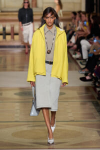 Runway model wearing a bright yellow jacket over a gray dress, with a long beaded necklace, belt, white heels, and a gray handbag.