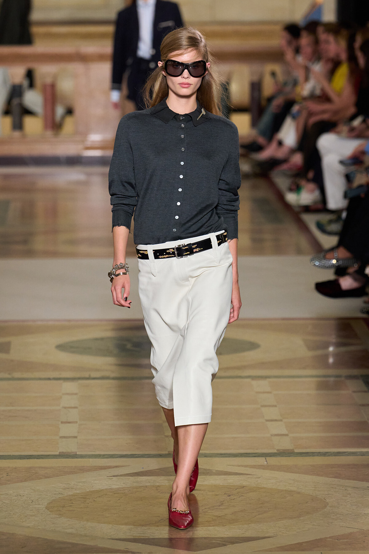 Model walking a runway in a dark gray button-down sweater, white cropped trousers, black belt, red flats, and oversized sunglasses.
