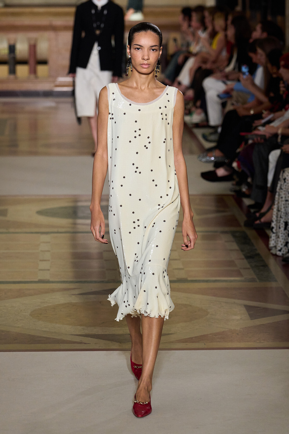 Model walking a runway in a cream sleeveless polka-dot dress with a ruffled hem and red flats.