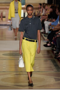 Model walks the runway wearing a dark polo tucked into bright yellow cropped trousers with a black belt, carrying a white handbag.