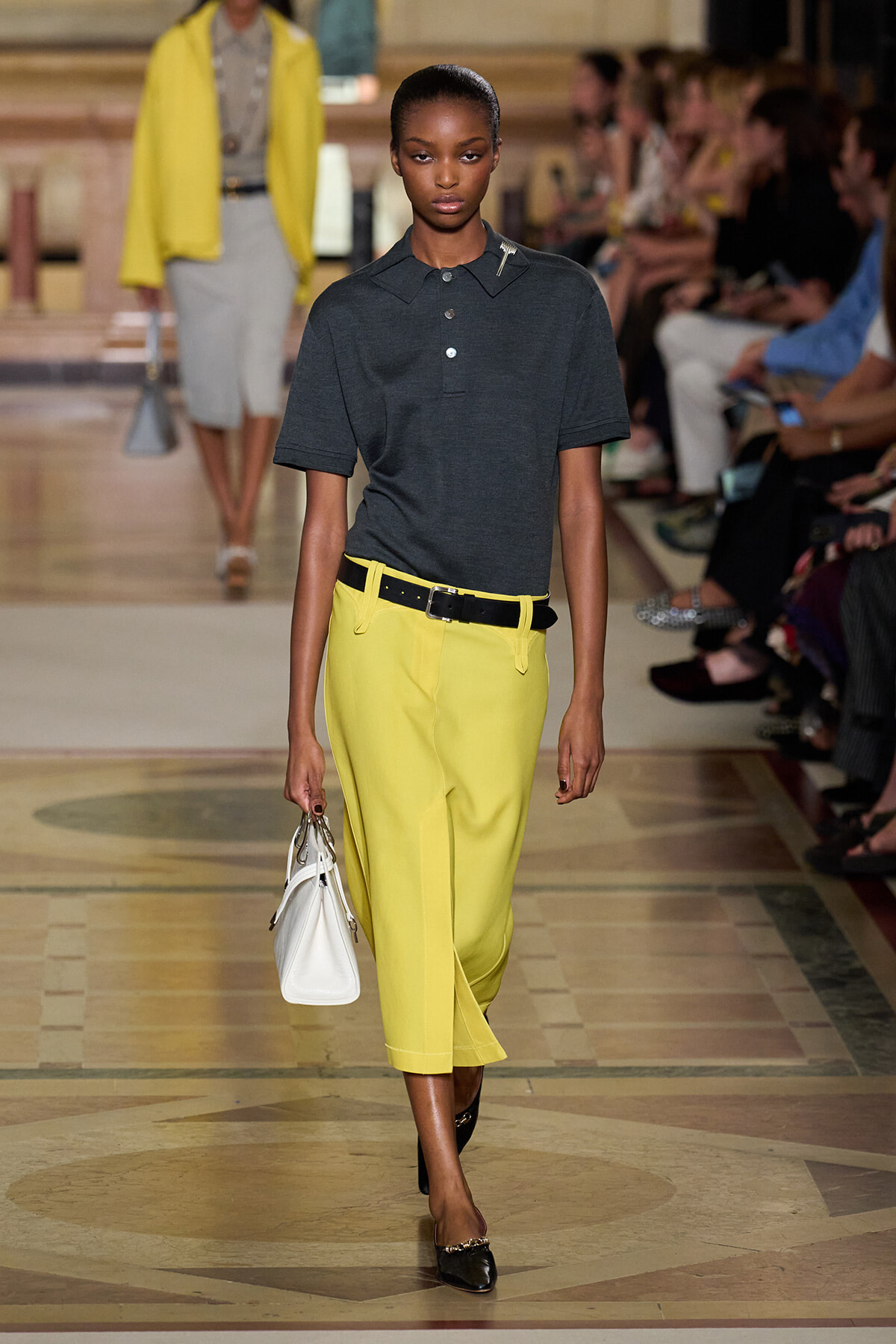 Model walks the runway wearing a dark polo tucked into bright yellow cropped trousers with a black belt, carrying a white handbag.