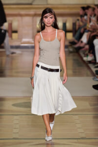 Model walks a runway in a beige ribbed tank top with side ties and a white midi skirt, accessorized with a black belt and white heels.