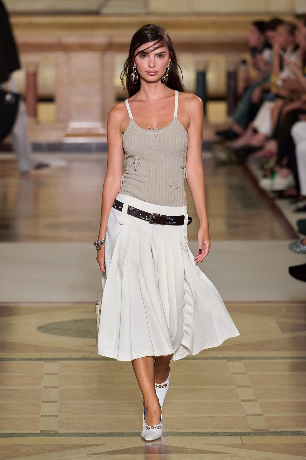 Model walks a runway in a beige ribbed tank top with side ties and a white midi skirt, accessorized with a black belt and white heels.