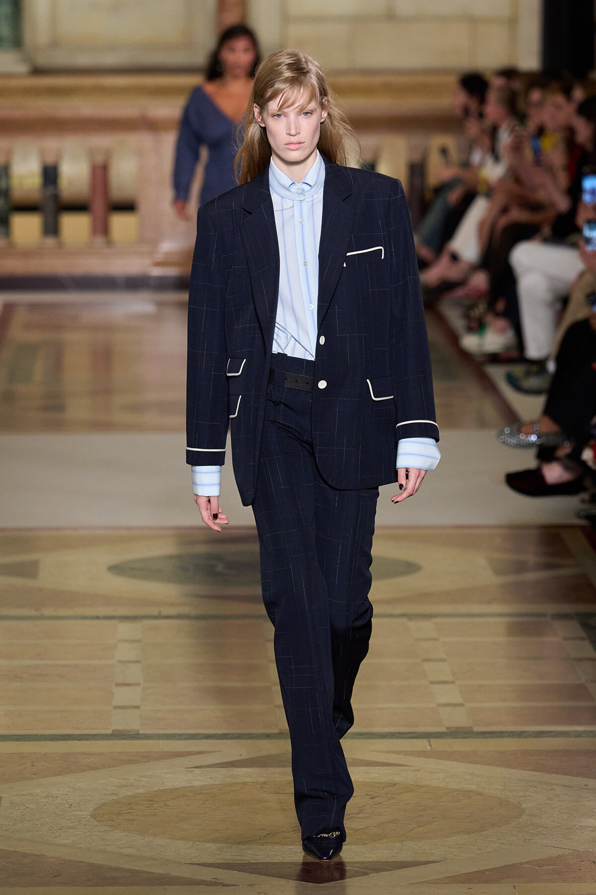 Model walks the runway in a navy pinstripe suit with a light blue shirt and white piping details on the pockets and cuffs.