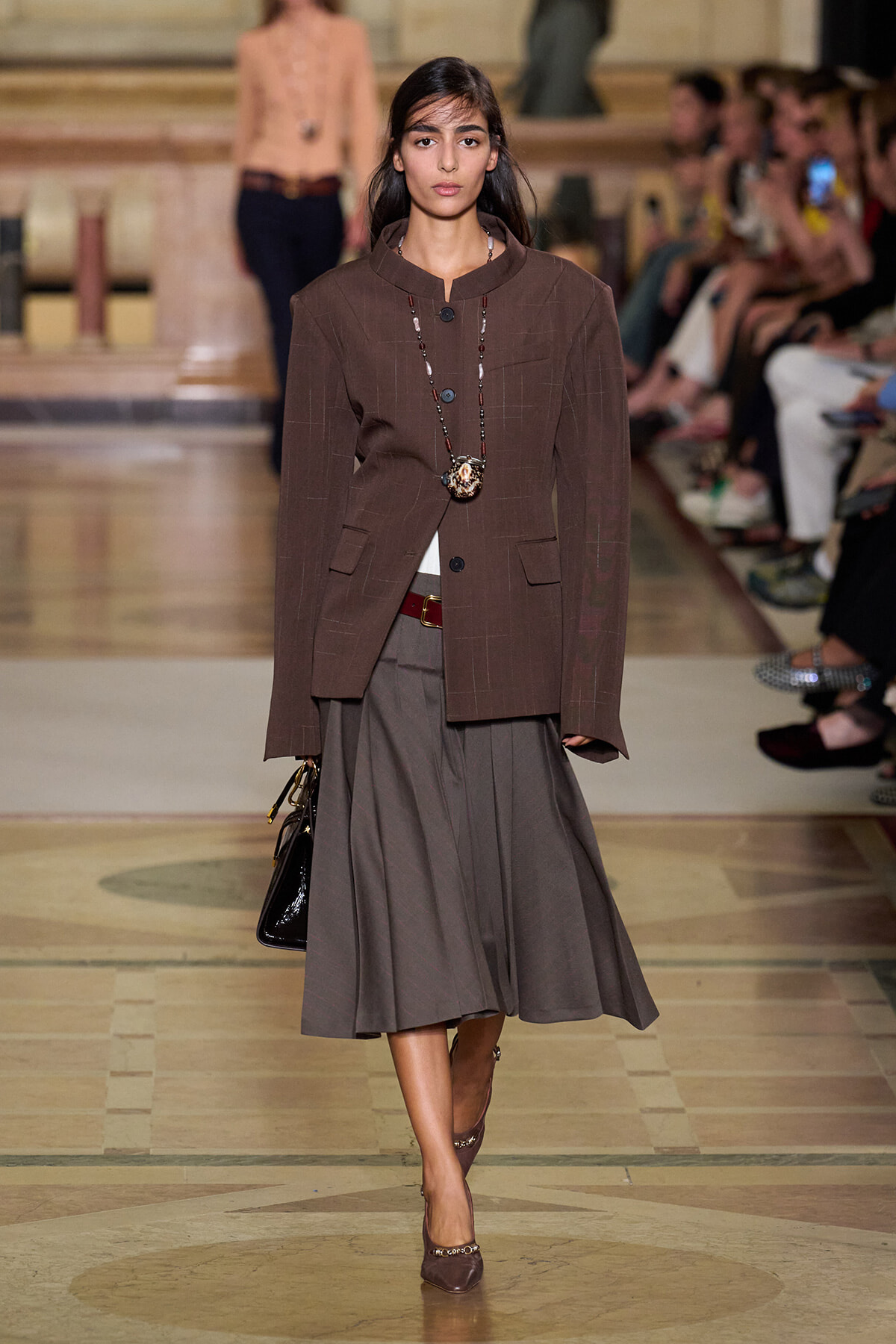 Model on a fashion runway wearing a brown tailored blazer over a brown top and a gray midi skirt, accessorized with a long beaded necklace and a black handbag.