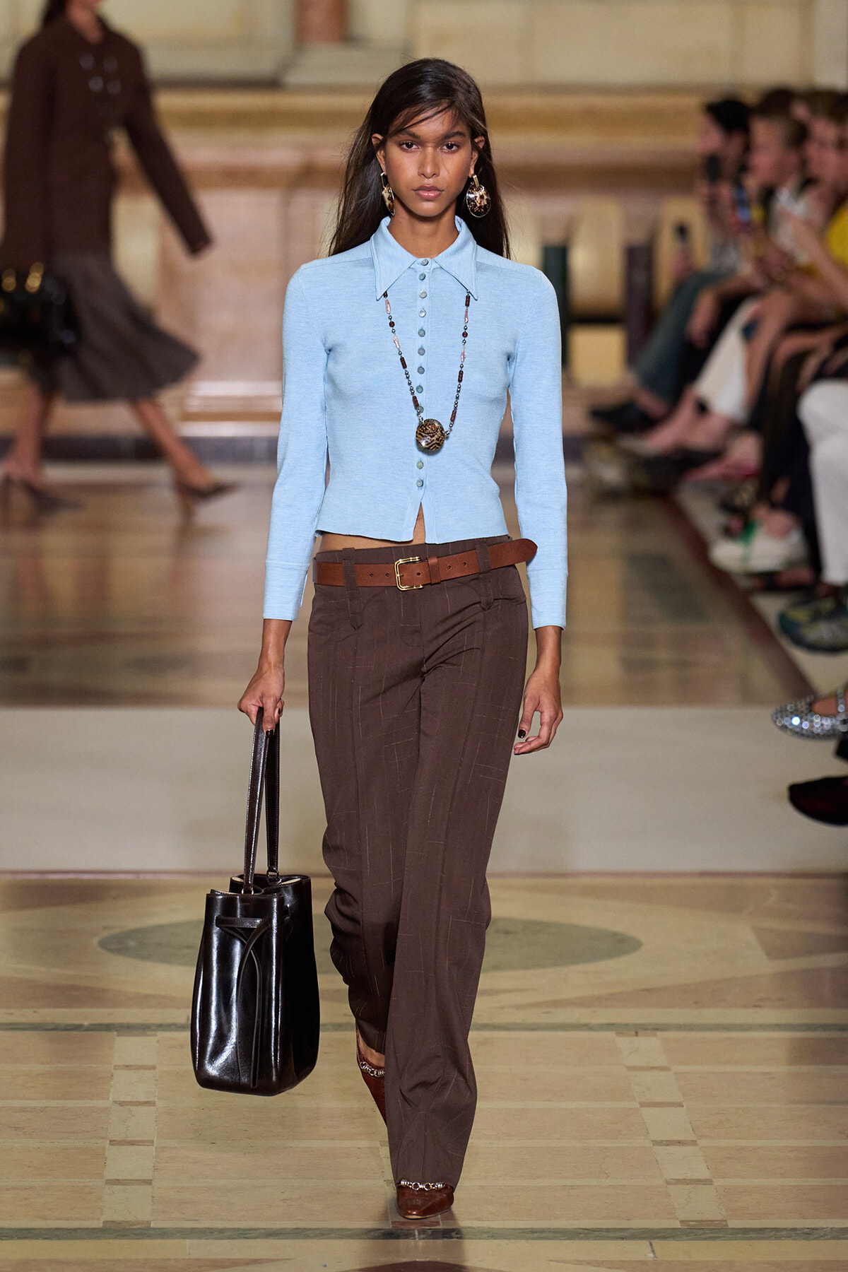 Female model walking a runway in a light blue fitted cardigan, brown pinstripe trousers with a brown belt, statement necklace, gold earrings, and a large black tote.