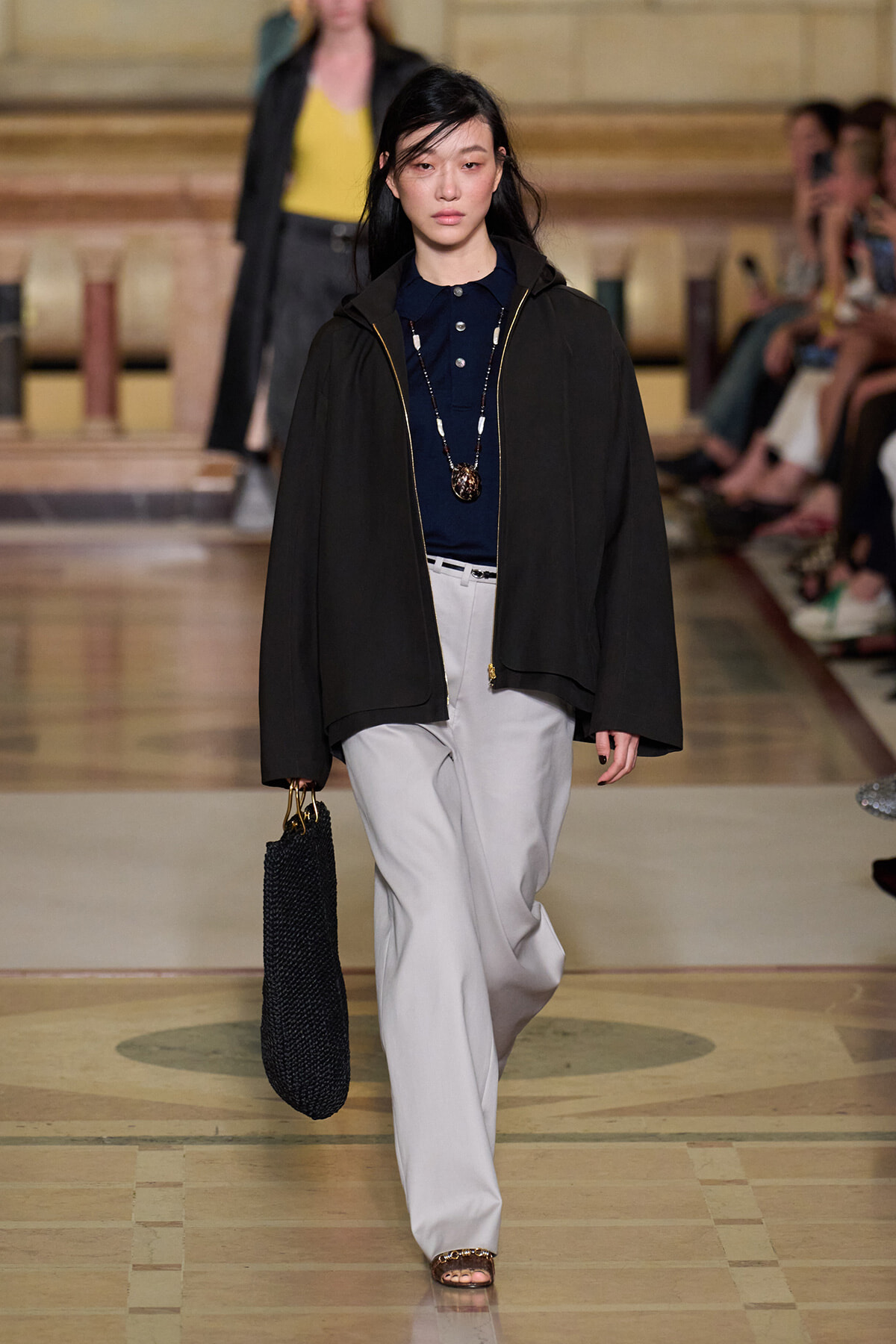 Model walks a fashion runway in a dark oversized jacket, navy polo, light gray wide-leg pants, and a black woven tote bag.