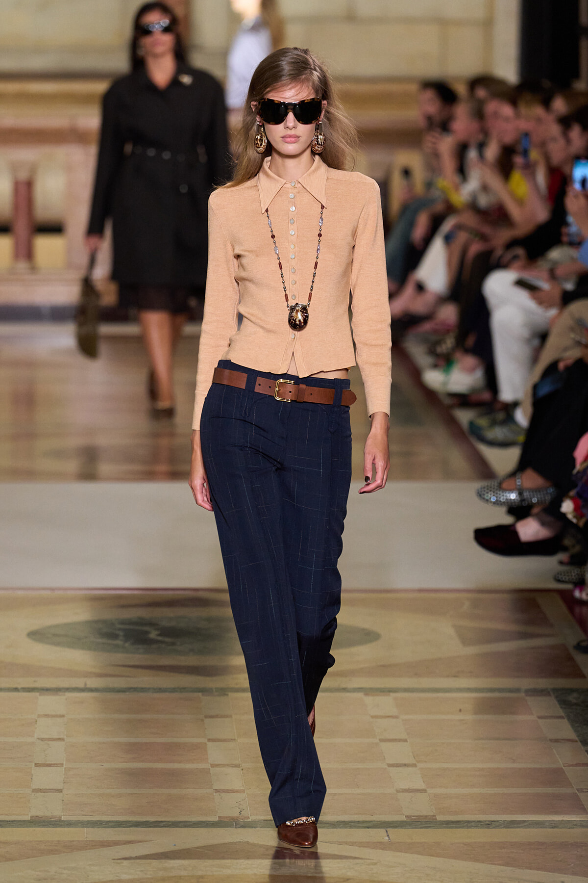 Model walks the runway in a beige buttoned cardigan with navy pinstriped trousers and a brown belt, accessorized with sunglasses and long beaded necklace.