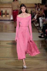Model walks the runway in a pink off-the-shoulder long-sleeve top with a matching flowing pink skirt; audience visible in background.