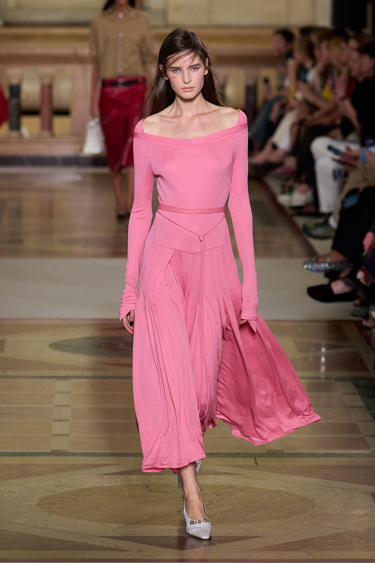 Model walks the runway in a pink off-the-shoulder long-sleeve top with a matching flowing pink skirt; audience visible in background.