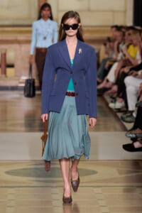 Model walking runway in a blue pinstripe blazer over a teal top and teal pleated skirt, brown heels, oversized sunglasses, and brown handbag on a fashion show floor.