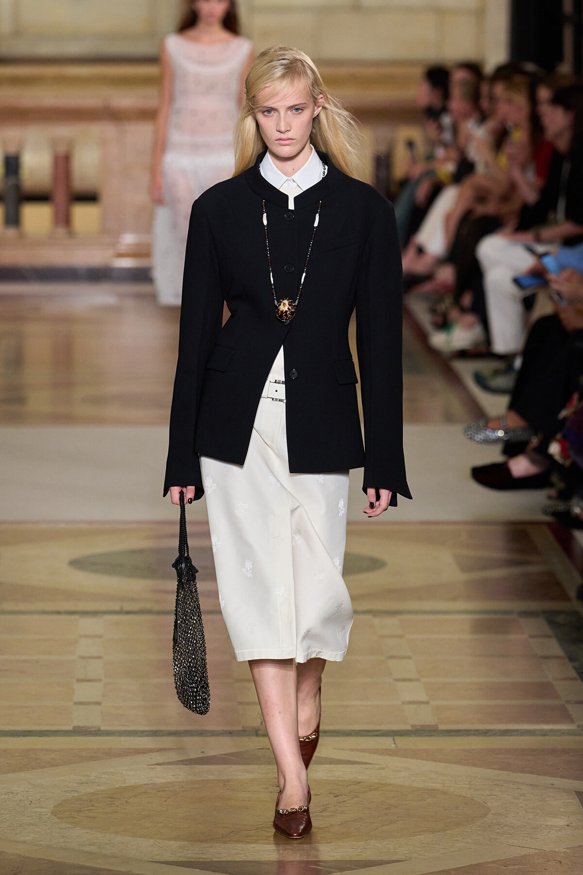 Model walking runway in a black blazer over a white shirt and ivory midi skirt, accessorized with a long beaded necklace and a black beaded bag.