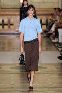 Model walking runway in a light blue polo tucked into brown wide-leg cropped pants with a black belt, carrying a small black handbag and wearing navy heels.