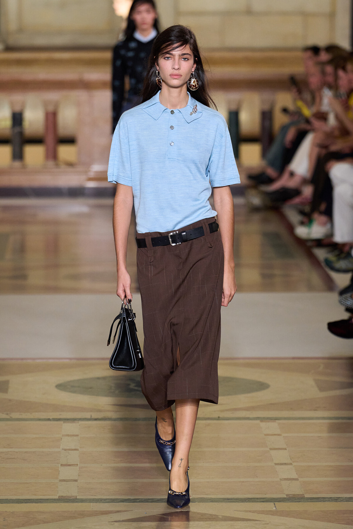 Model walking runway in a light blue polo tucked into brown wide-leg cropped pants with a black belt, carrying a small black handbag and wearing navy heels.
