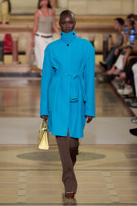 Model walks the runway in a bright turquoise belted coat with a high collar, brown trousers, and brown shoes, carrying a pale yellow handbag.