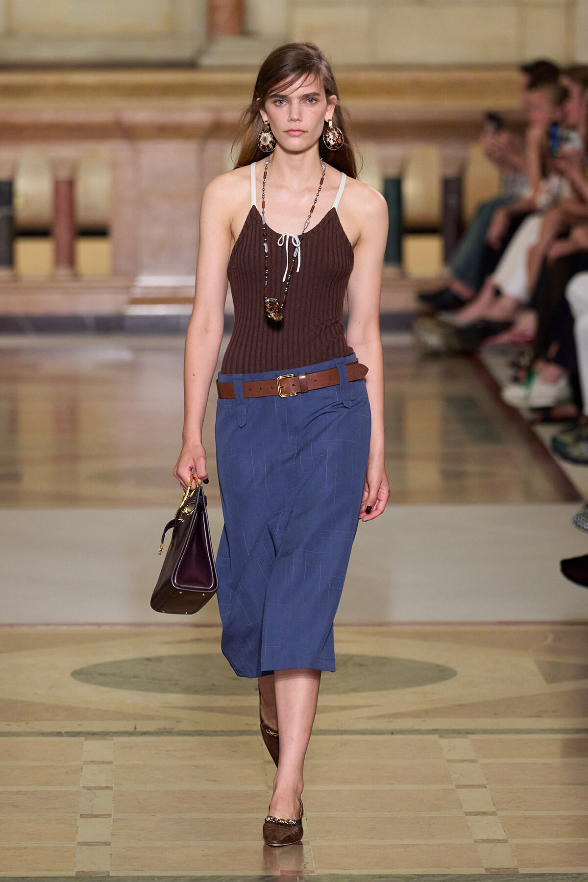 Model walking a runway in a brown ribbed tank top, blue midi skirt, brown belt, and brown accessories while carrying a dark handbag, jewelry visible.