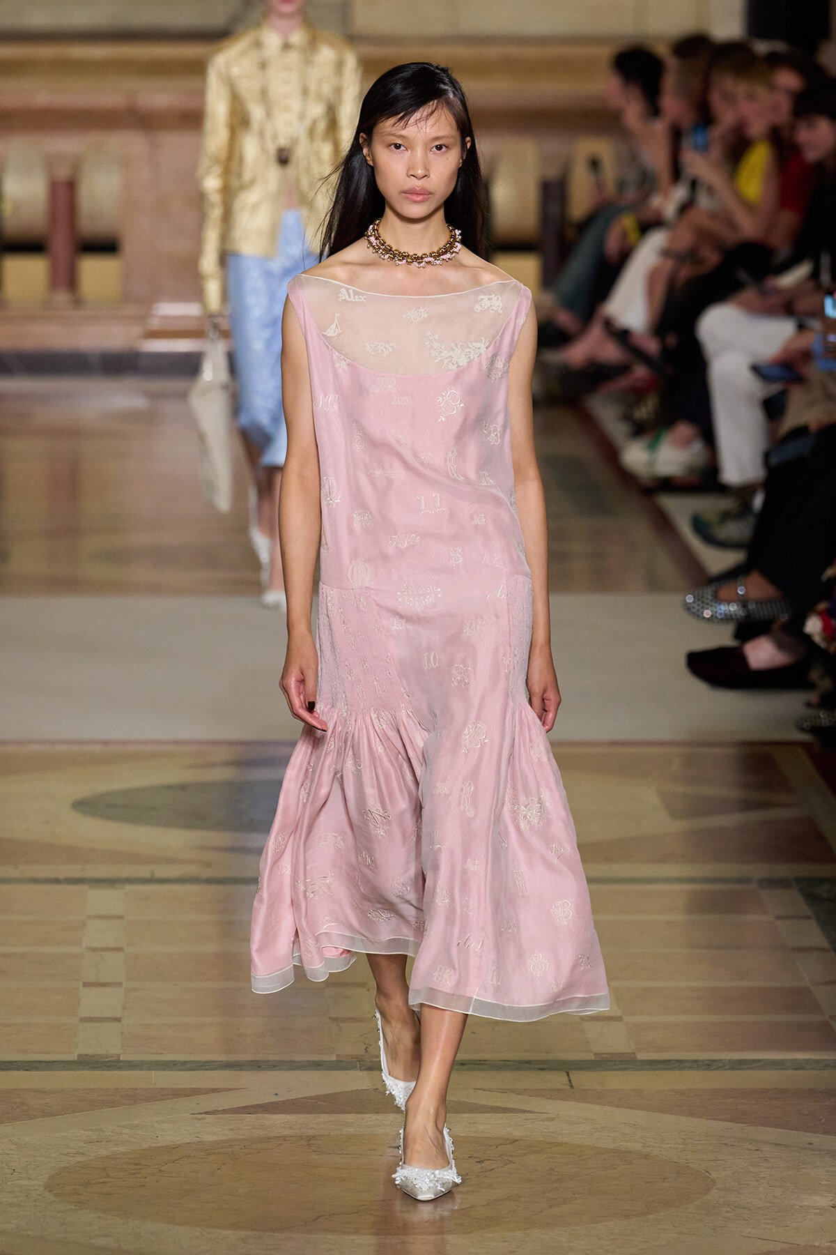 Model walking a fashion runway in a pale pink sleeveless dress with a sheer neckline and delicate embroidered motifs, wearing a pearl necklace and white embellished heels.