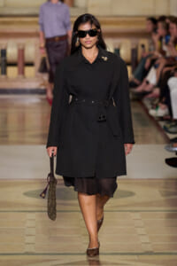 Model walks down a fashion runway in a long black belted coat, oversized sunglasses, and gold earrings, carrying a chain bag.