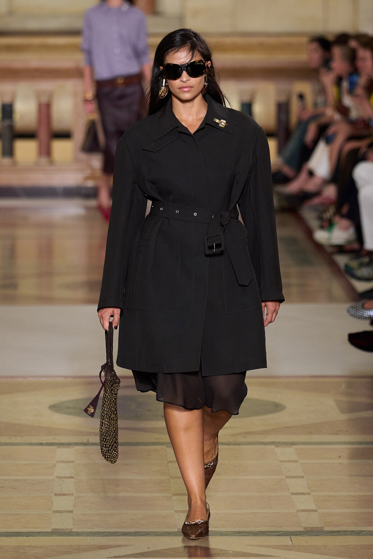 Model walks down a fashion runway in a long black belted coat, oversized sunglasses, and gold earrings, carrying a chain bag.