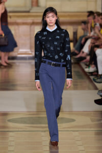 Model walking a runway in a navy embroidered sweater, light blue collar, and blue pinstripe trousers with a black belt and brown shoes.