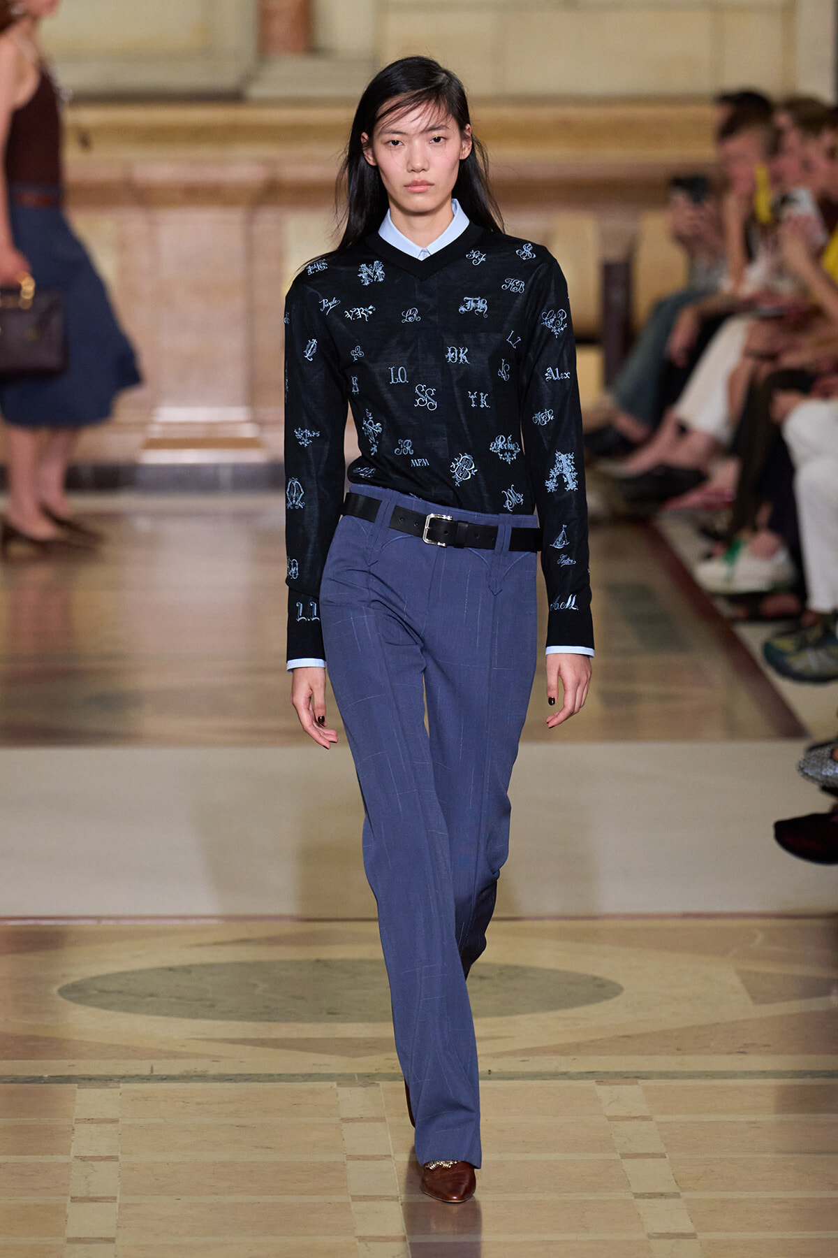 Model walking a runway in a navy embroidered sweater, light blue collar, and blue pinstripe trousers with a black belt and brown shoes.
