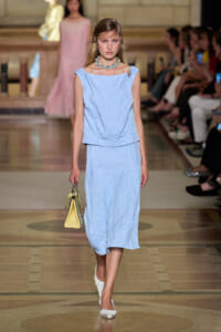 Model walks the runway in a light blue, crinkled sleeveless top and midi skirt, with a turquoise necklace and a yellow handbag.
