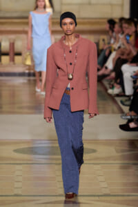 Model walking the runway in a dusty-rose blazer, blue crinkled pants, black headscarf, and long beaded necklace with brown shoes.