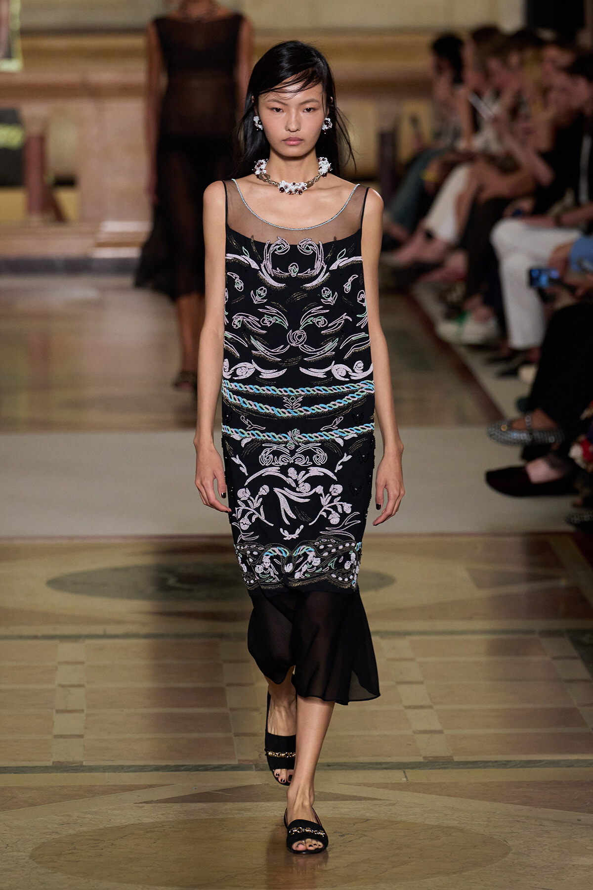 Model walking a runway in a black sleeveless dress with sheer neckline and intricate white and turquoise embroidery, accessorized with matching jewelry.