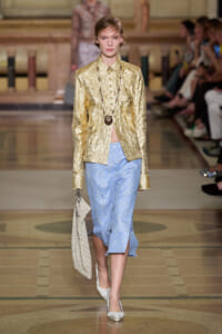 Runway model in a crinkled metallic gold shirt with a light blue midi skirt, layered beaded necklace, and a cream crochet handbag.