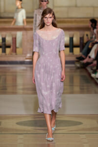 Model walking the runway in a lavender midi dress with sheer, embroidered neckline and short sleeves, on a marble-patterned fashion show floor.