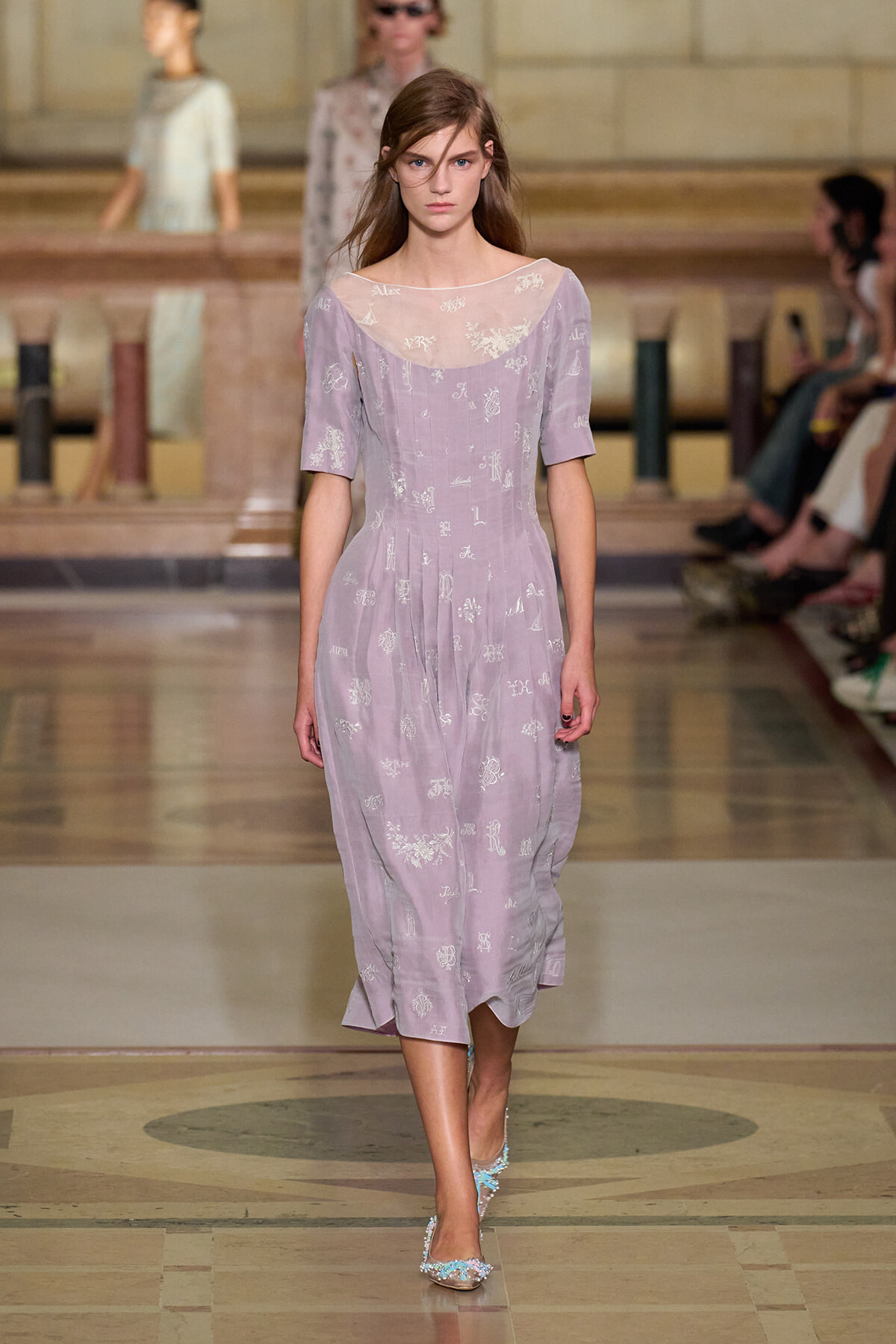 Model walking the runway in a lavender midi dress with sheer, embroidered neckline and short sleeves, on a marble-patterned fashion show floor.
