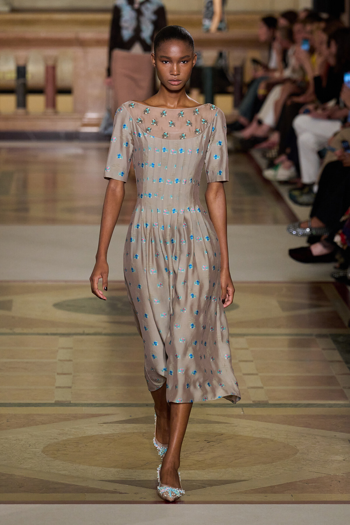 Model walks a beige midi dress with blue floral embroidery down the runway, paired with sparkling pointed-toe heels.