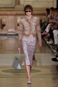 Model walking a runway in a taupe embroidered cardigan and lavender patterned midi skirt, with sunglasses, pearl necklace, and a gray handbag.