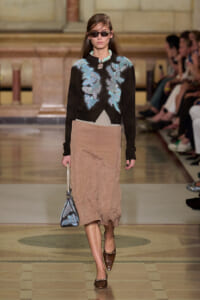 Model walks runway in black cropped cardigan with blue floral embroidery, taupe bias-cut skirt, brown heels, and blue patterned handbag while wearing sunglasses at a fashion show.