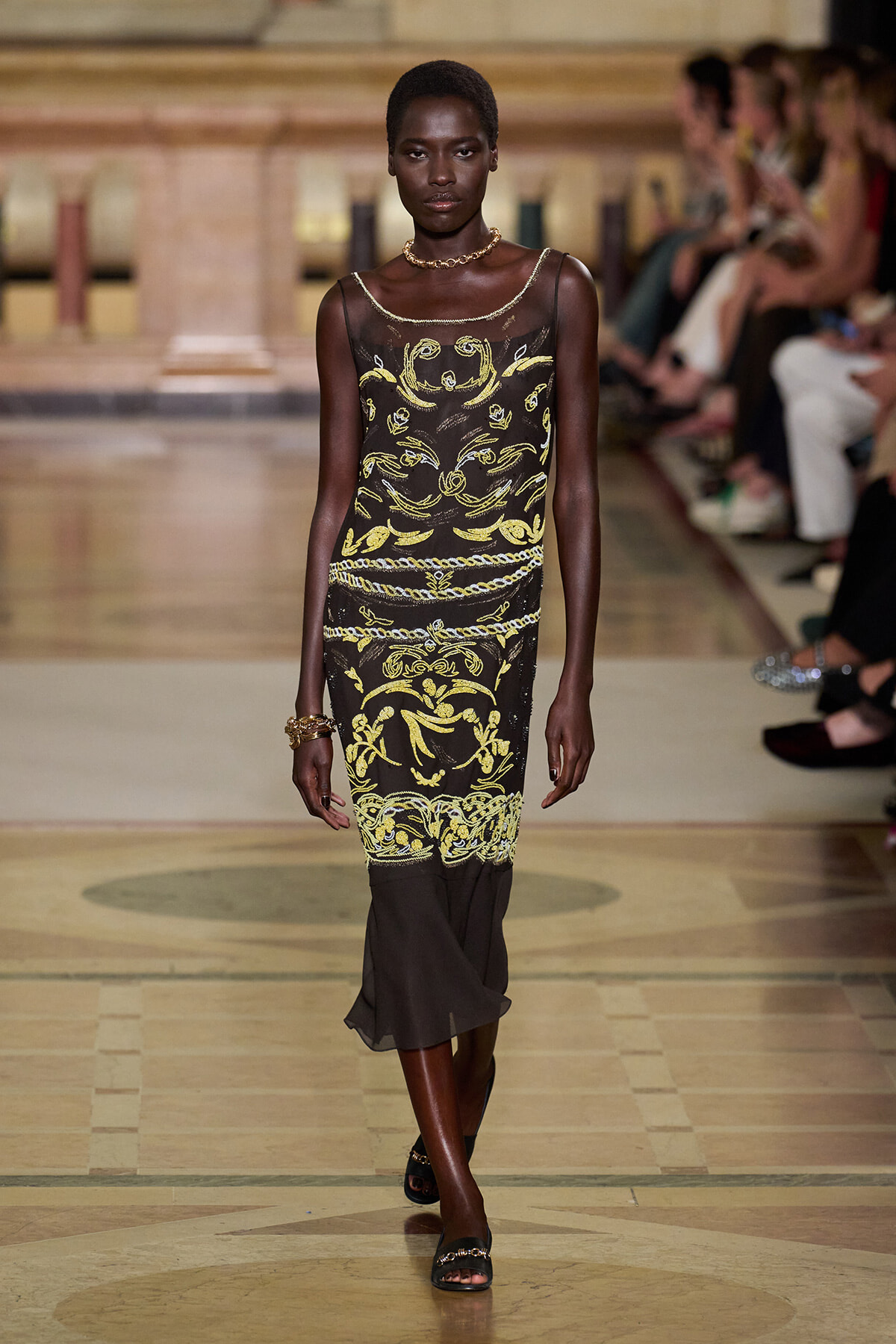 Runway model wearing a sheer black dress with gold ornate embroidery, accessorized with a necklace and bracelets as she walks the catwalk in a fashion show.