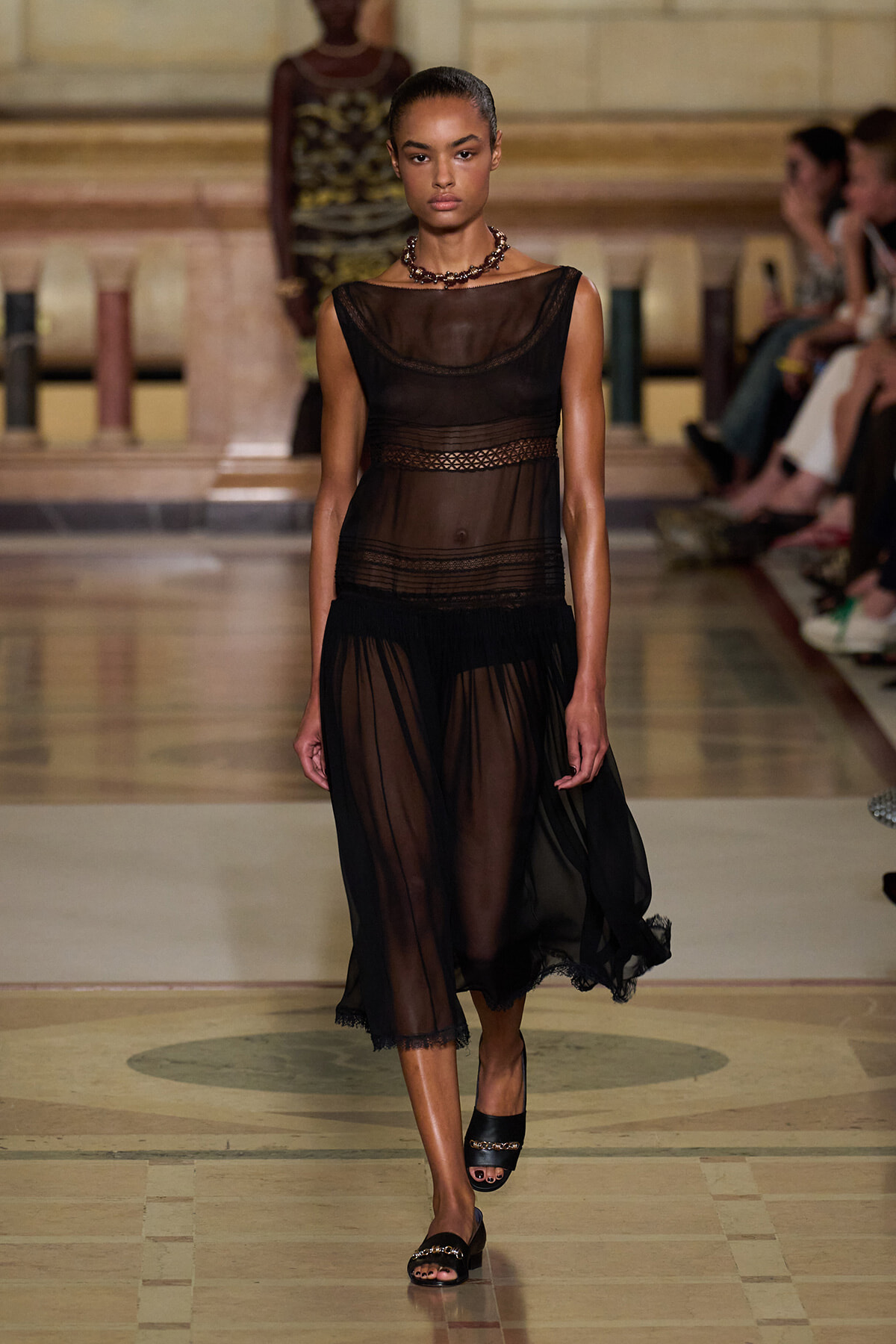 Model walks the runway in a sheer black midi dress with mesh panels and lace trim, paired with black sandals and a chunky necklace.