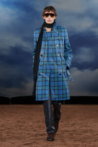 Model wearing a blue plaid double-breasted coat, black scarf, sunglasses, and brown boots walking on a sandy runway with a cloudy sky backdrop.
