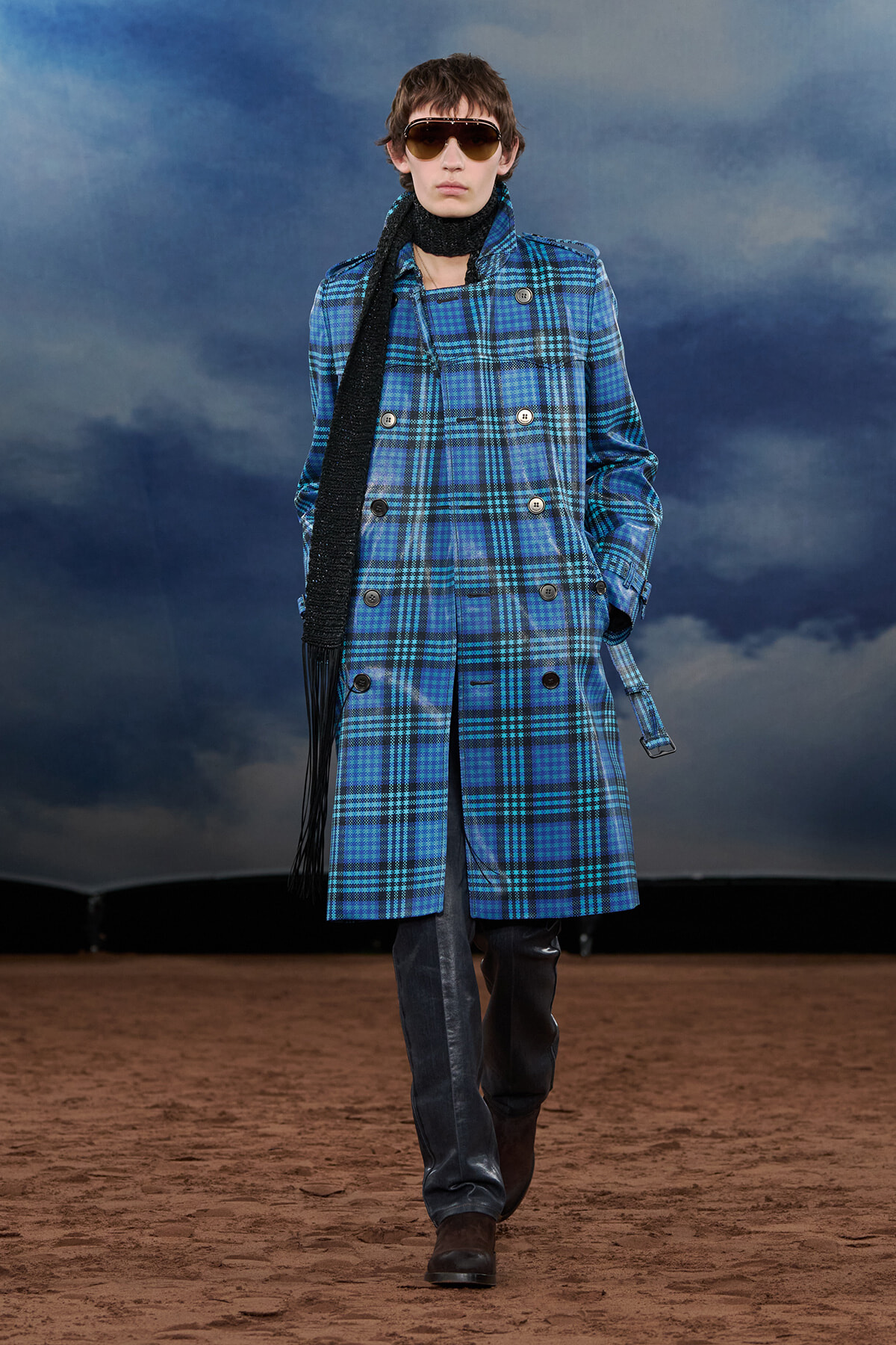 Model wearing a blue plaid double-breasted coat, black scarf, sunglasses, and brown boots walking on a sandy runway with a cloudy sky backdrop.