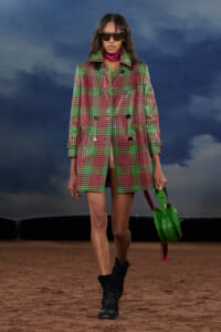 Model walks a runway in a green and pink plaid coat, black boots, oversized sunglasses, and a bright green handbag.