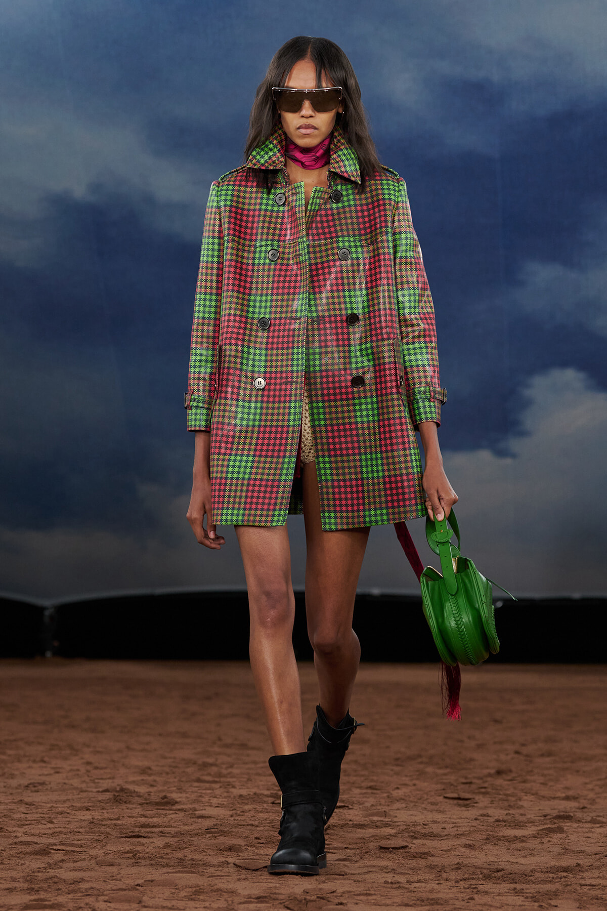 Model walks a runway in a green and pink plaid coat, black boots, oversized sunglasses, and a bright green handbag.