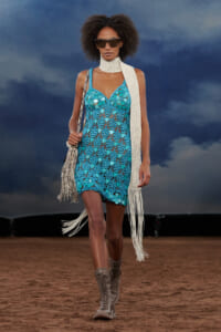 Model walking a fashion runway in a turquoise crochet mini dress with a long white fringe scarf and oversized sunglasses, carrying a fringed bag.