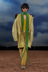 Male model walking runway in a oversized yellow plaid coat, mustard top, tan trousers, green scarf, and sunglasses against a stormy sky backdrop.