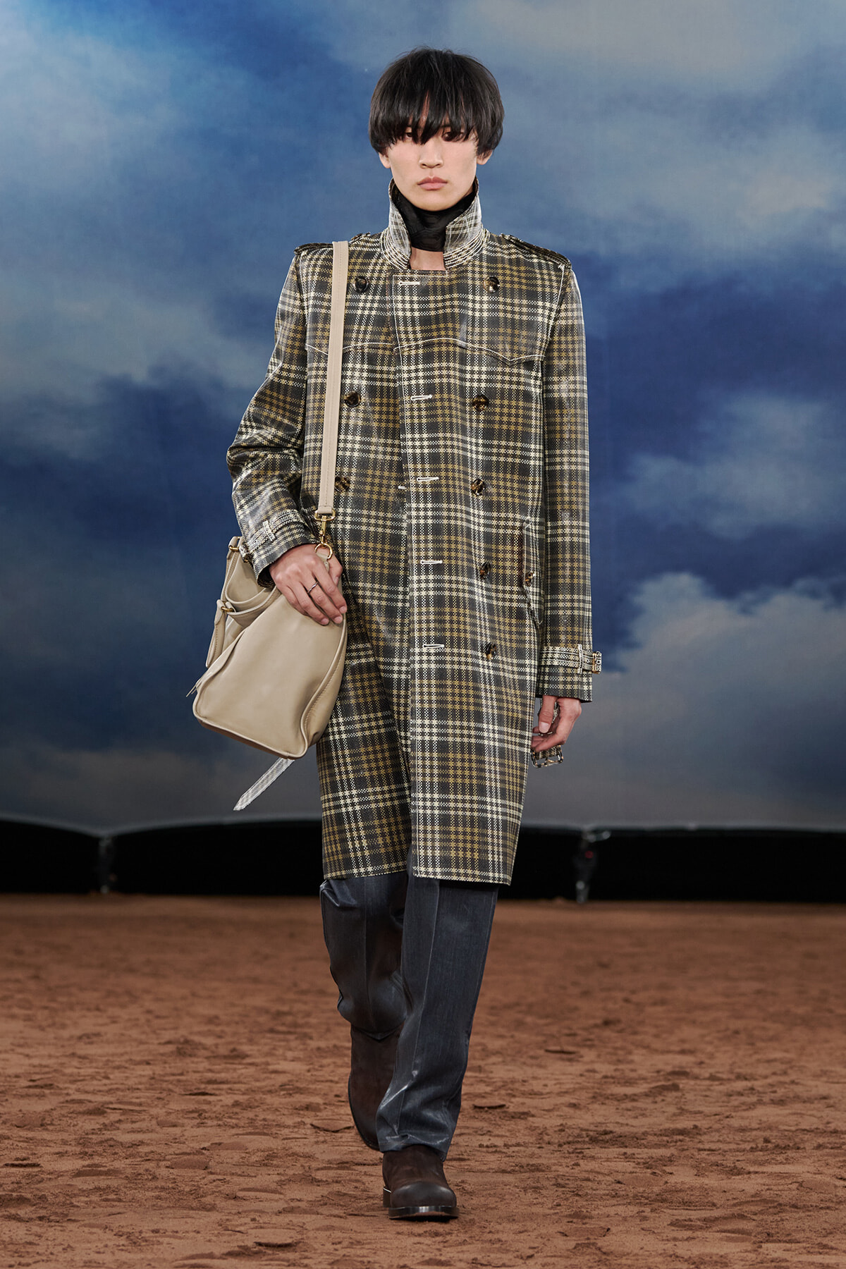 Model walks a runway wearing a long plaid double-breasted coat in beige, gray, and brown, with a black turtleneck, dark jeans, brown boots, and a beige shoulder bag.