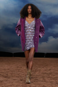 Model walking a fashion show runway in a purple oversized jacket over a lilac lace dress, tan boots, against a blue cloudy backdrop.